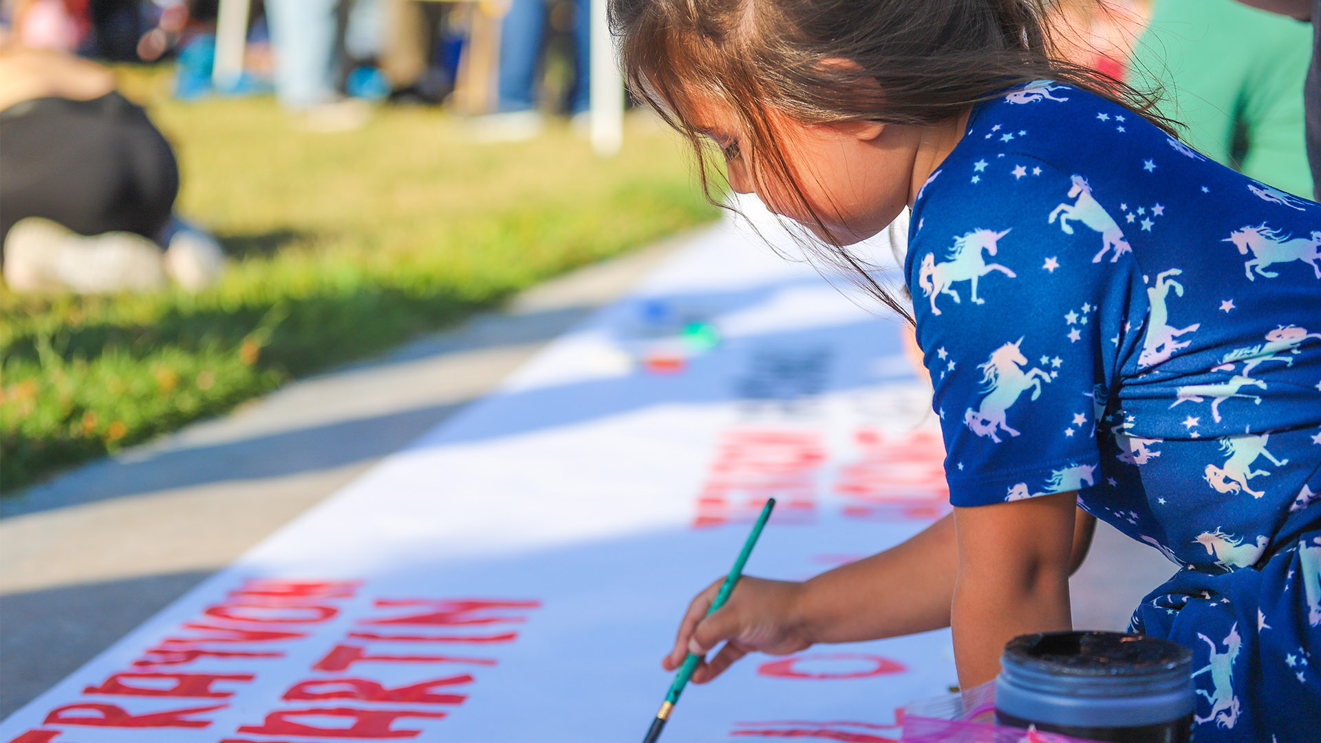 5 year old girl painting name of African American life lost to police on banner in park.